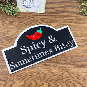 May include: A black and white sign featuring the text "Spicy & Sometimes Bitey" and a red chili pepper illustration. The sign has a white border and two small holes for hanging. It rests on a wooden surface.