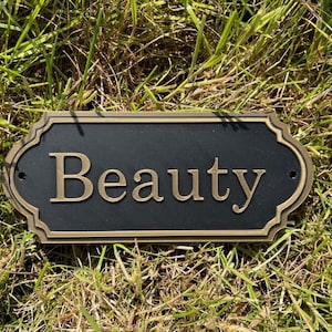 May include: A black metal sign with a gold border and the word "Beauty" in gold lettering.