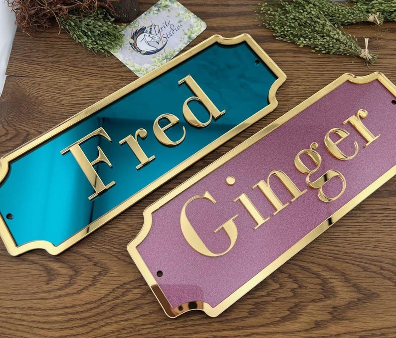 May include: Two rectangular nameplates with gold trim. One is teal with "Fred" in gold letters, the other is pink with "Ginger" in gold letters. Both have a glossy finish and are on a wooden surface.
