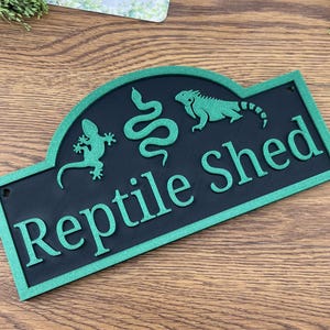 May include: A black and green sign reading "Reptile Shed". The sign features green silhouettes of a lizard, a snake, and an iguana. The sign has a decorative arched top and is rectangular. The sign is made of wood.
