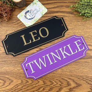 May include: Two personalised horse stable signs. The first sign is black with gold lettering that reads "LEO". The second sign is purple with silver lettering that reads "TWINKLE".