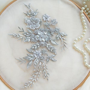 Pode incluir: Aplique floral prateado com contas e lantejoulas brancas. O aplique está sobre um fundo branco.