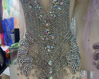 May include: A sheer fabric garment with a decorative design made of iridescent rhinestones. The design features a V-neckline and a pattern of swirls and curves.