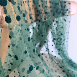 Può includere: Tessuto verde trasparente con pois verde scuro. Il tessuto è drappeggiato su un bastone di legno.