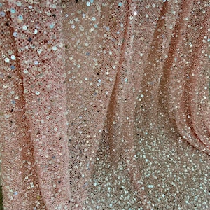 May include: A light pink fabric featuring a dense overlay of small, circular, reflective sequins. The material has a mesh-like base structure, creating a textured appearance with shimmering highlights across the surface of the draped textile.