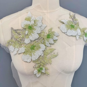 Può includere: Applicazione floreale con perline con fiori bianchi e verde chiaro e dettagli argentati. L'applicazione è attaccata a un tessuto trasparente bianco sporco. Questo ornamento decorativo è ideale per aggiungere un tocco di eleganza a capi o accessori.