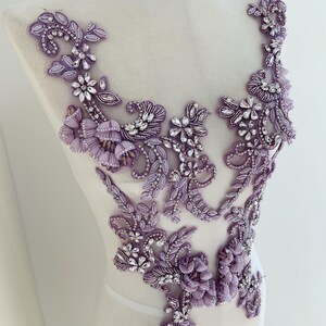 Può includere: Applicazione floreale lavanda con intricato lavoro di perline e scintillanti dettagli in cristallo. Il design presenta fiori, foglie e motivi a spirale, ideale per abbellire capi di abbigliamento o accessori. L'applicazione è esposta su un manichino.