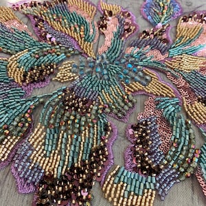 May include: An intricate floral appliqué featuring a dense arrangement of metallic gold, teal, and iridescent purple glass beads and sequins. The design is meticulously hand-stitched onto a sheer, fine-mesh fabric base with purple thread borders.
