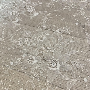 Könnte beinhalten: Nahaufnahme eines weißen Spitzenstoffs mit Blumen- und Blättermustern. Die zarte Spitze ist mit kleinen, verstreuten Pailletten verziert. Der Stoff wird wahrscheinlich für Brautmode, Kleider oder andere dekorative Projekte verwendet.