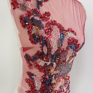 以下が含まれることがあります： 複雑な花の刺繍が施された、透明なバーガンディ色の生地のクローズアップ。赤、ピンク、青、金のスパンコールとビーズが組み合わされ、テクスチャード加工された装飾的な要素を作り出しています。この生地は、アパレルやクラフトプロジェクトに使用される可能性があります。