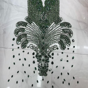 Puede incluir: Un diseño de adorno de cristal verde en forma de lágrima. El diseño está hecho de pequeños cristales facetados que se cosen juntos para crear un patrón decorativo.