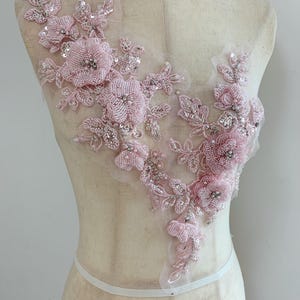 Puede incluir: Una aplicación floral de color rosa claro con intrincados abalorios y lentejuelas sobre una base de malla transparente. El diseño consta de flores tridimensionales, hojas delicadas y patrones de enredadera en forma asimétrica.