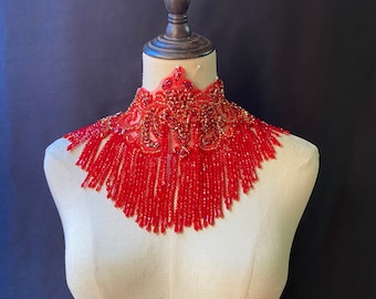 May include: A red beaded choker necklace with a floral design and fringe. The necklace is displayed on a white mannequin.