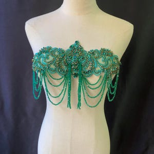 Pode incluir: Uma aplicação bordada com contas verde-azuis e douradas com fios de contas penduradas. O design ornamentado apresenta bordados intrincados e embelezamentos brilhantes. A aplicação é exibida em um manequim.