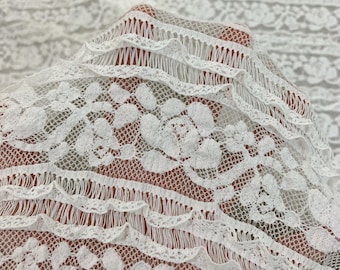 May include: Close-up of white lace fabric with a floral pattern. The delicate lace features intricate designs and a scalloped edge. The fabric is soft and airy, suitable for dresses, skirts, or craft projects.
