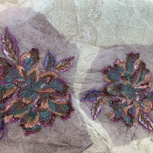 Può includere: Due applicazioni floreali con intricate perline. I disegni presentano un mix di colori, tra cui verde acqua, viola e oro, con accenti di paillettes. Le applicazioni sono su un tessuto trasparente, lilla chiaro, ideali per decorare.