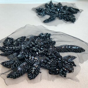 May include: Two black floral-shaped appliqués crafted from sheer black mesh fabric. Each piece is heavily embellished with black beads, sequins, and faceted crystals arranged in a dense, intricate pattern that creates a textured, reflective surface.