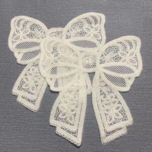 May include: Two white lace bows with intricate floral designs. The bows are layered on top of each other and have a delicate, feminine look.