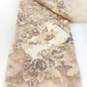 May include: Beige lace fabric with 3D floral embroidery. The fabric features detailed, raised floral designs in shades of beige, cream, and mauve, with beaded accents. The delicate lace is folded, revealing the intricate patterns and textures of the embellishments.