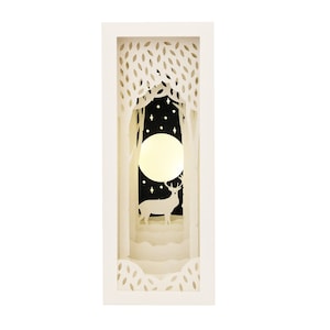 May include: A white papercraft 3D scene depicting a deer standing in a forest under a full moon. The scene is illuminated from behind, creating a glowing effect.