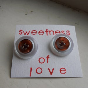 Könnte beinhalten: Zwei weiße Knöpfe mit braunen Mittelteilen und einem Vierloch-Design. Die Knöpfe befinden sich auf einer weißen Karte mit dem Text "sweetness of love" in Rot.