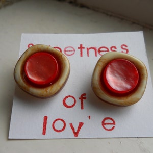 Könnte beinhalten: Zwei Vintage-Knöpfe mit roter Mitte und cremefarbenem und braunem Außenring. Die Knöpfe befinden sich auf einem weißen Papier mit den rot gedruckten Worten "sweetness of love".