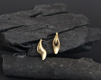 May include: A pair of gold-toned earrings with a unique, abstract design. The earrings are shaped like a teardrop with a cutout in the center. They are displayed on a black, textured surface.