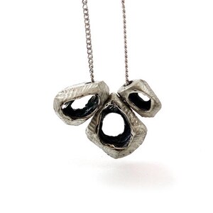 May include: A silver-toned necklace featuring three textured, open-center pendants. Each pendant has a unique, irregular shape and is suspended from a delicate chain. The interior of each pendant is dark, creating a contrast with the silver exterior.