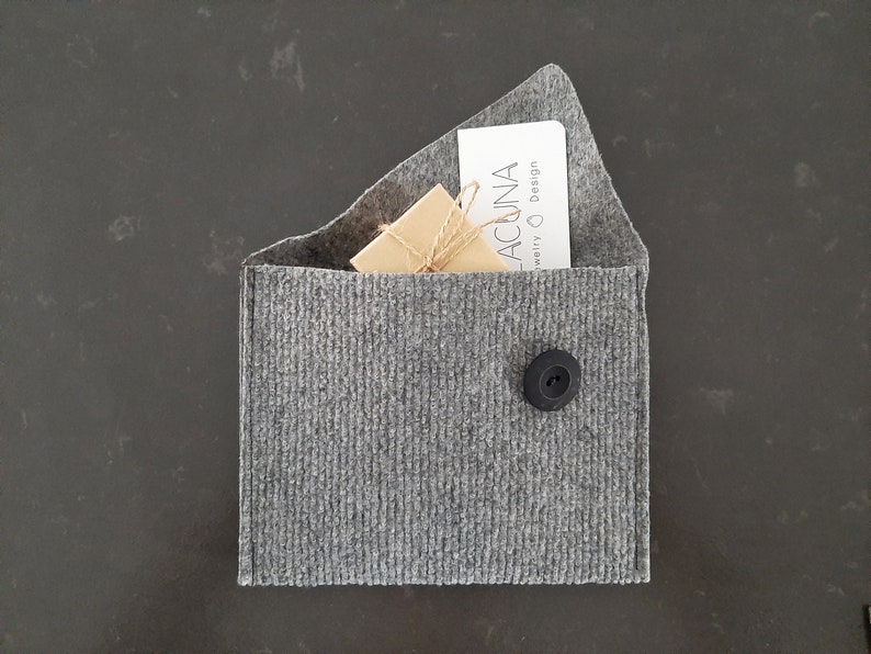 May include: A gray felt envelope with a black button closure. Inside the envelope is a small brown box tied with twine and a white card with the text "ACUNA Jewelry & Design".