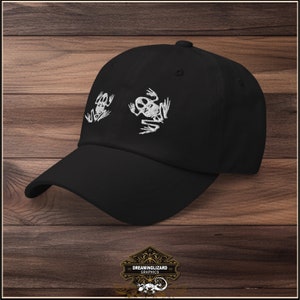 Puede incluir: Gorra de béisbol negra con diseños de ranas esqueleto bordadas en blanco. La gorra tiene una visera curvada y una correa ajustable.