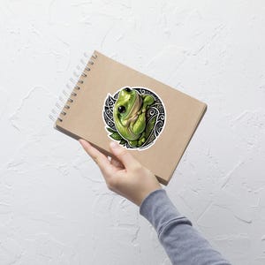 May include: A spiral-bound notebook with a tan cover featuring a circular sticker of a green frog. The frog is depicted with large black eyes and a light green body, set against a background of intricate black and white swirling patterns and leaves.