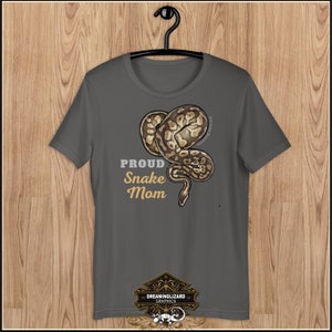 Op de afbeelding: Een donkergrijs T-shirt met een bruin en wit slangenontwerp en de tekst "Proud Snake Mom". Het shirt heeft ook een klein logo onderaan dat "Dreaminglizard Graphics" zegt.