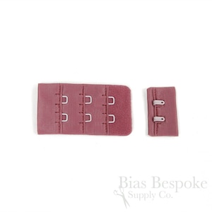May include: A dusty rose bra hook and eye closure set consisting of two pieces. The larger piece features three columns of two metal hooks, while the smaller piece contains two metal eyes. The fabric is a textured, matte material in a muted pink.