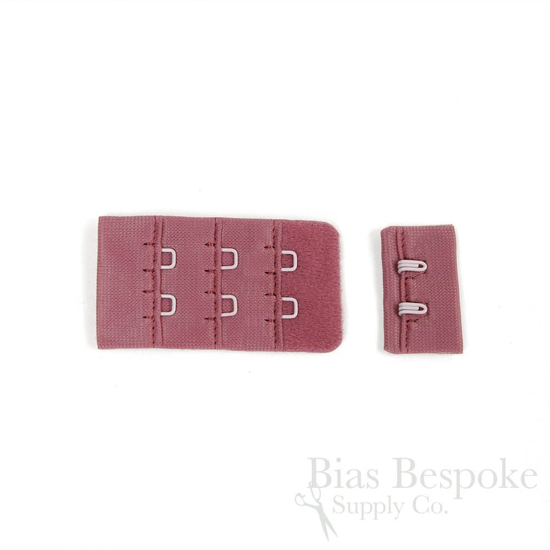 May include: A dusty rose bra hook and eye closure set consisting of two pieces. The larger piece features three columns of two metal hooks, while the smaller piece contains two metal eyes. The fabric is a textured, matte material in a muted pink.