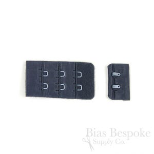 May include: A dark navy blue bra extender featuring three rows of metal hooks and eyes on a rectangular fabric base, accompanied by a smaller matching fabric piece with two metal loops, designed for adjusting the fit of undergarment bands.