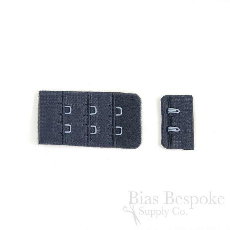 May include: A dark navy blue bra extender featuring three rows of metal hooks and eyes on a rectangular fabric base, accompanied by a smaller matching fabric piece with two metal loops, designed for adjusting the fit of undergarment bands.
