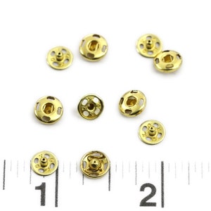 以下が含まれることがあります： 金色の金属製のスナップファスナー10個のクローズアップ。ファスナーは白い表面にランダムなパターンで配置されています。画像の下部に定規が見えます。