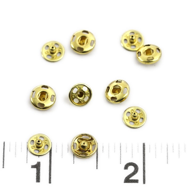 以下が含まれることがあります： 金色の金属製のスナップファスナー10個のクローズアップ。ファスナーは白い表面にランダムなパターンで配置されています。画像の下部に定規が見えます。