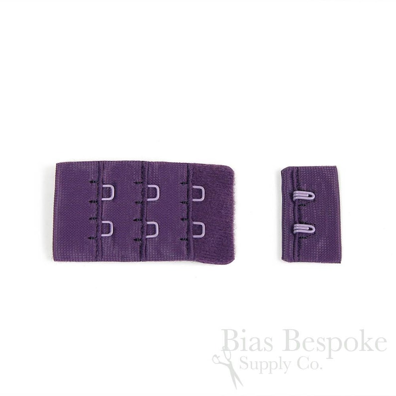 May include: A purple bra extender featuring three columns of hook-and-eye closures on the left piece and two rows of eyes on the right piece. The fabric has a textured finish and the metal hardware is coated in a matching purple color.