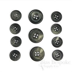 Puede incluir: Conjunto de diez botones de cuerno negros y verdes con cuatro agujeros cada uno. Los botones están dispuestos en tres filas sobre un fondo blanco.