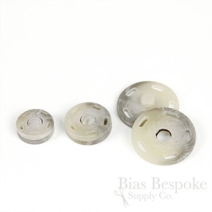 May include: Three white and gray plastic snap fasteners of different sizes. The fasteners have a swirl pattern and four holes around the edge.