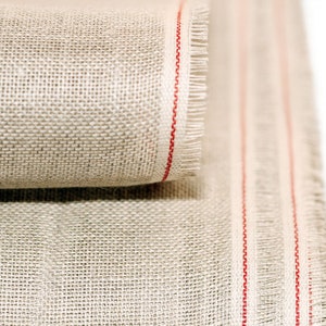 Puede incluir: Un rollo de tela de lino natural con rayas rojas a lo largo de los bordes. La tela está tejida con un tejido apretado y uniforme y tiene una textura ligeramente rugosa.