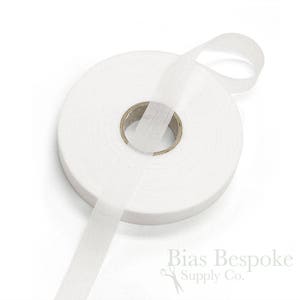 May include: A roll of white bias tape, a type of fabric used for finishing edges of garments. The tape is wound around a cardboard spool and a length of the tape is extending from the spool.