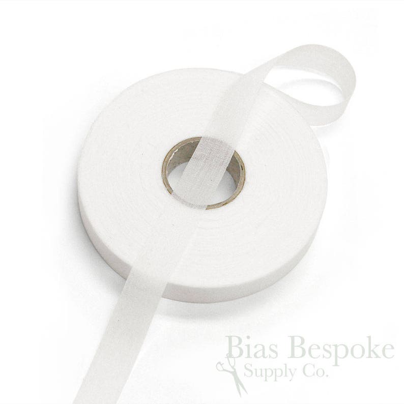 May include: A roll of white bias tape, a type of fabric used for finishing edges of garments. The tape is wound around a cardboard spool and a length of the tape is extending from the spool.