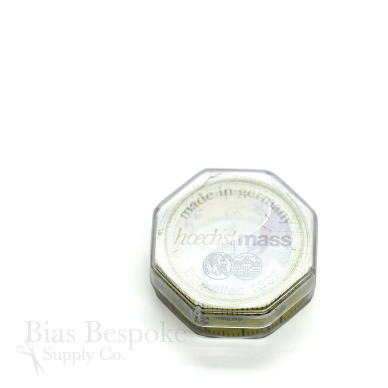 May include: A clear, octagonal tape measure case with a white tape inside. The tape measure is marked with both inches and centimeters. The text "made in germany" and "hoechstmass" are visible on the tape measure case.