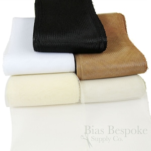 May include: A stack of four rolls of tulle fabric in black, white, beige, and brown. The fabric is a fine mesh with a soft, delicate texture. Bias Bespoke Supply Co. is printed on the bottom roll.