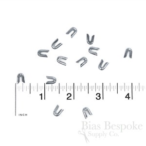 May include: A collection of small, silver-colored metal staples scattered on a white surface. A ruler with inch measurements is visible, with the staples ranging in size. The staples are U-shaped and likely used for crafting or sewing projects.