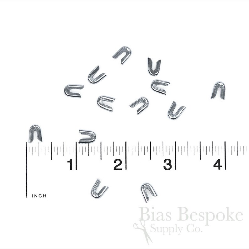 May include: A collection of small, silver-colored metal staples scattered on a white surface. A ruler with inch measurements is visible, with the staples ranging in size. The staples are U-shaped and likely used for crafting or sewing projects.