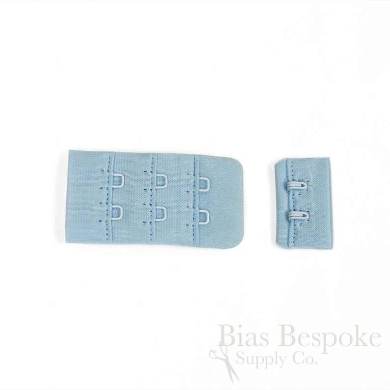 May include: A light blue bra hook and eye closure set featuring a wide three-column, two-row hook panel and a corresponding two-row eye panel. The fabric components are finished with matching light blue stitching and metal hardware components.