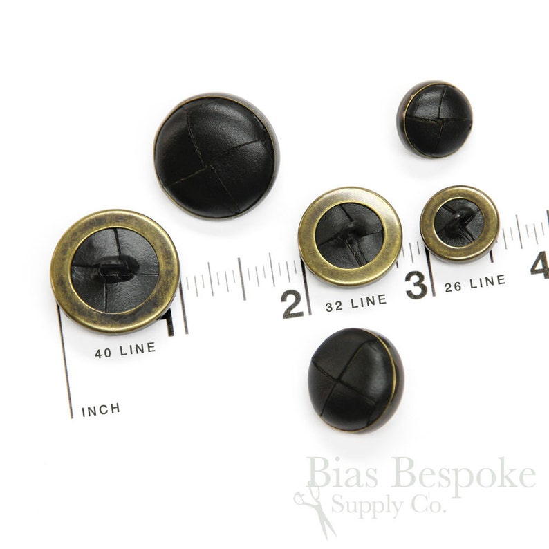 May include: Five black leather buttons with a brass rim. The buttons are different sizes and are arranged on a white surface with a ruler.